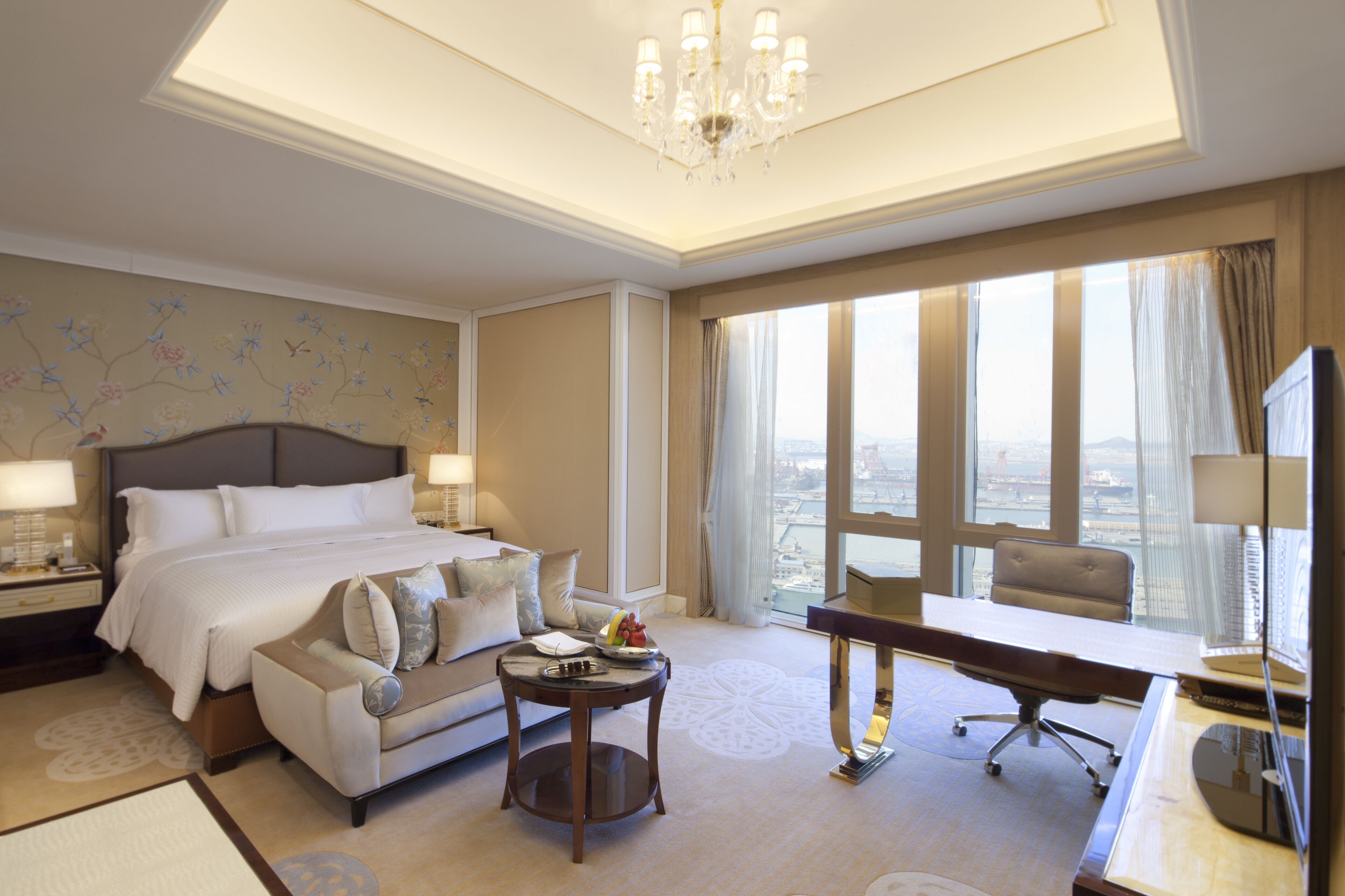 Une chambre d’hôtel équipée d’un lit, d’un bureau, d’une chaise, d’un canapé et offrant une vue sur la ville grâce à de grandes fenêtres.