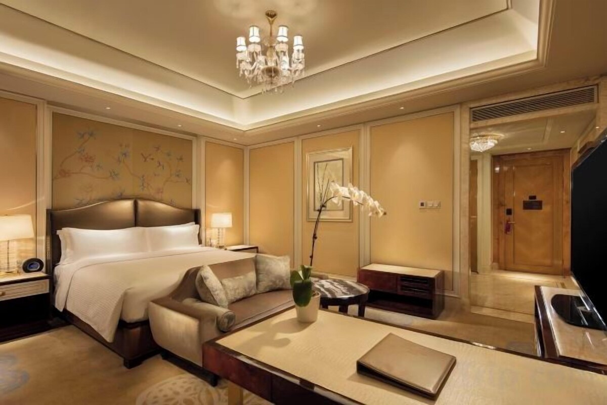 A hotel room with a large bed, a desk, a chair, a small table, and a decorative plant.