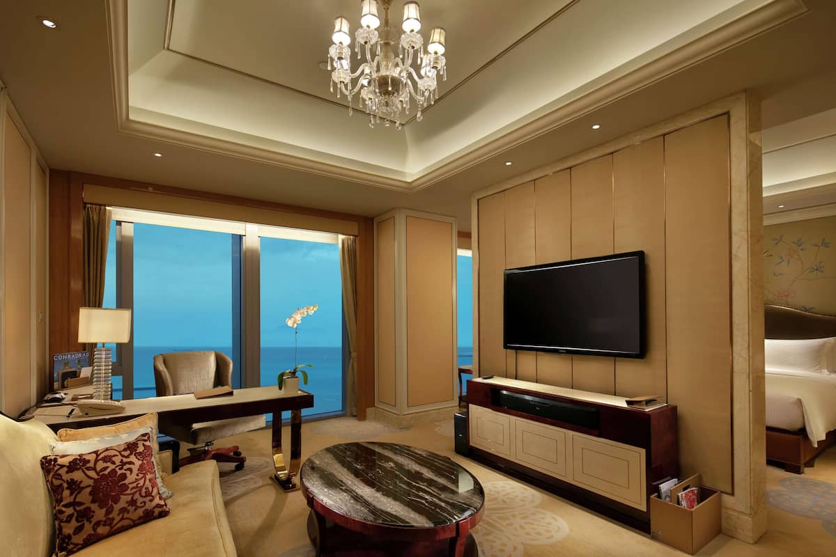 Ein Hotelzimmer mit großem Fernseher, einer Sitzecke, einem Couchtisch, einem Schreibtisch und Meerblick.