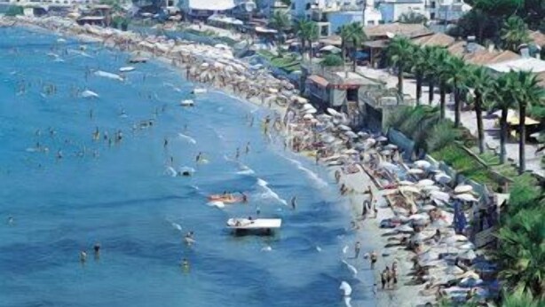 A crowded beach with people swimming and sunbathing, a boat, and buildings along the shore. - Hotel Melike (Kusadasi)