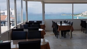 A restaurant with large windows offering a view of the sea and distant hills. Tables are arranged for dining. - Hotel Melike (Kusadasi)
