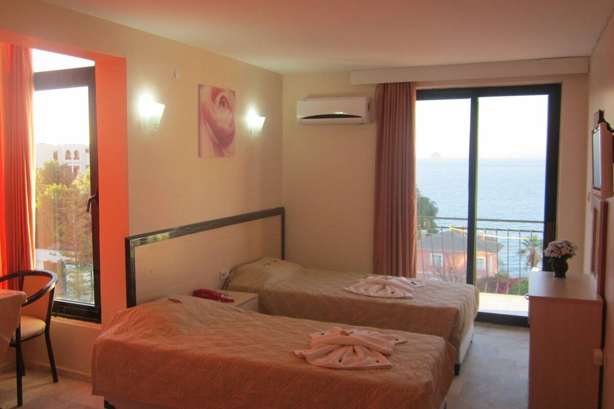 Habitación de hotel con dos camas, un escritorio y una silla. Cuenta con ventana con cortinas, unidad de aire acondicionado y vista al mar.