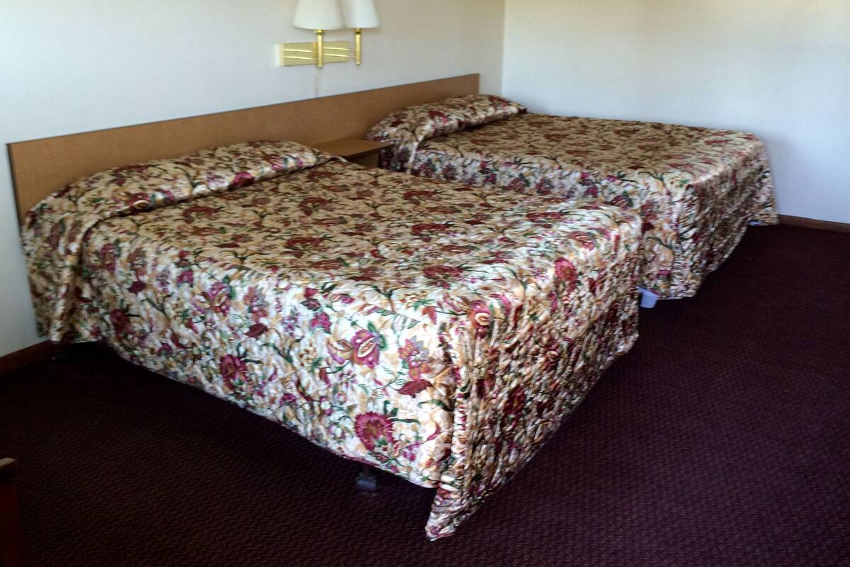 A hotel room with two beds, each with floral bedding, a wooden headboard, and a nightstand. There are two wall-mounted lamps above the beds.