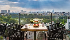 A table with a dessert, wine glasses, and a potted plant on a balcony overlooking a cityscape and greenery.