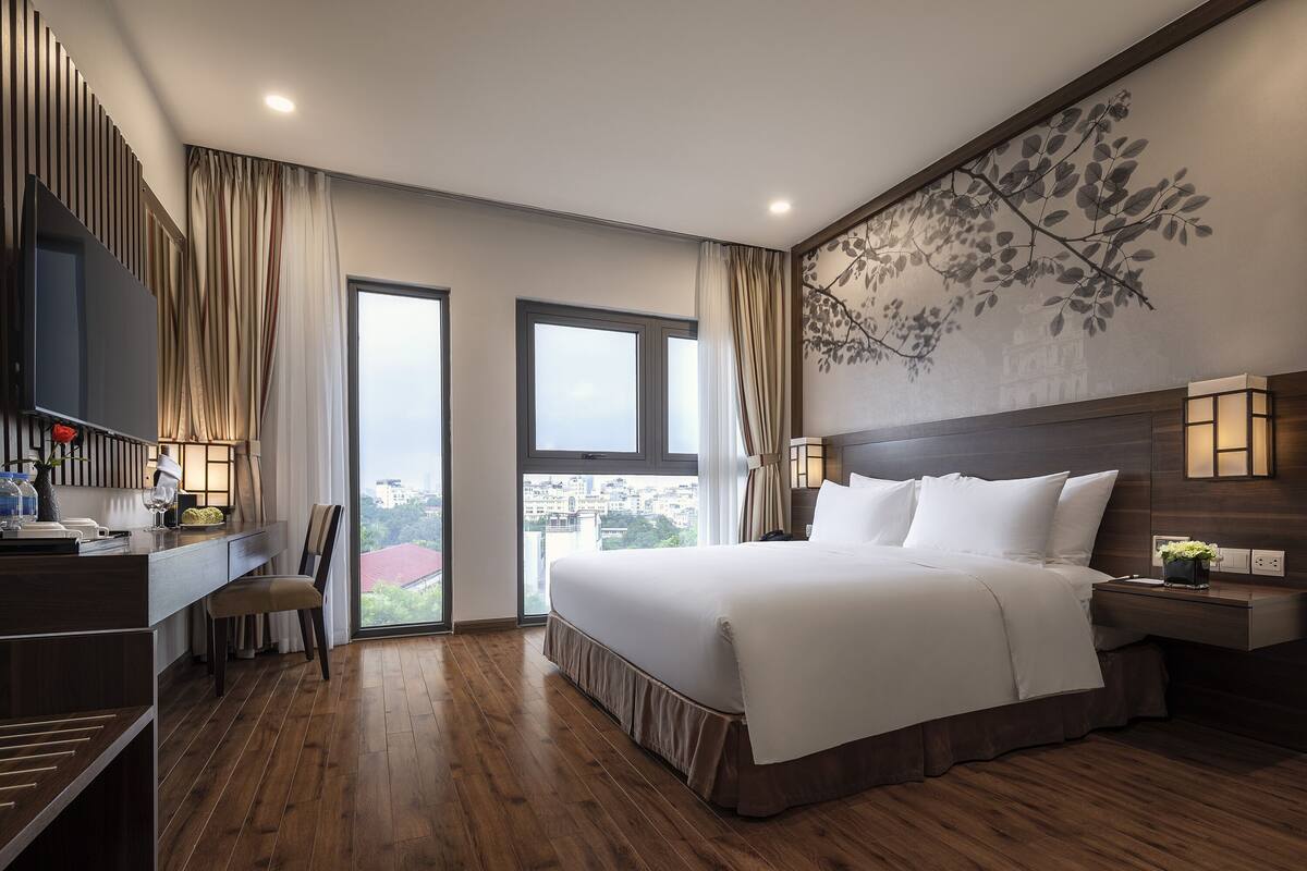 Ein modernes Hotelzimmer mit einem großen Bett, einem Schreibtisch mit Stuhl, einem Fernseher und Blick auf die Stadt durch große Fenster.