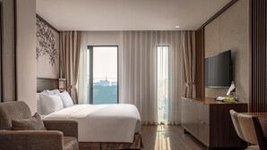 A modern hotel room with a large bed, a desk, a TV, and a view of the city.