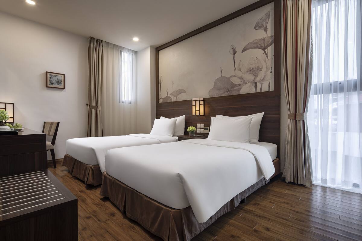 Ein Hotelzimmer mit zwei Betten, einem Schreibtisch, einem Sessel und einem Nachttisch. An der Wand hängt ein großes Blumenbild.