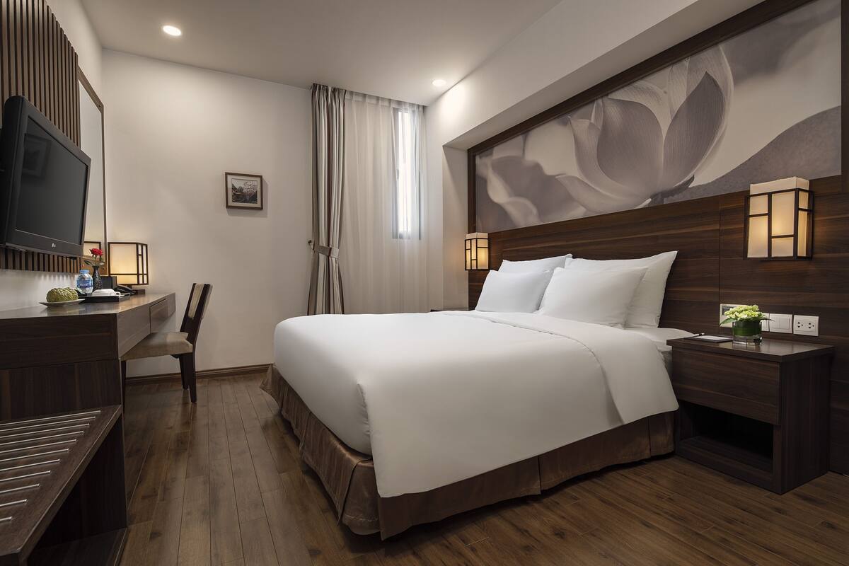 Ein modernes Hotelzimmer mit einem großen Bett, Holzboden, einem Schreibtisch mit Stuhl, einem Fernseher und einer Wandleuchte.