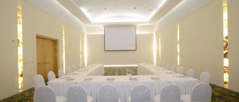 Un salón de conferencias con una larga mesa, sillas blancas y una pantalla de proyector.