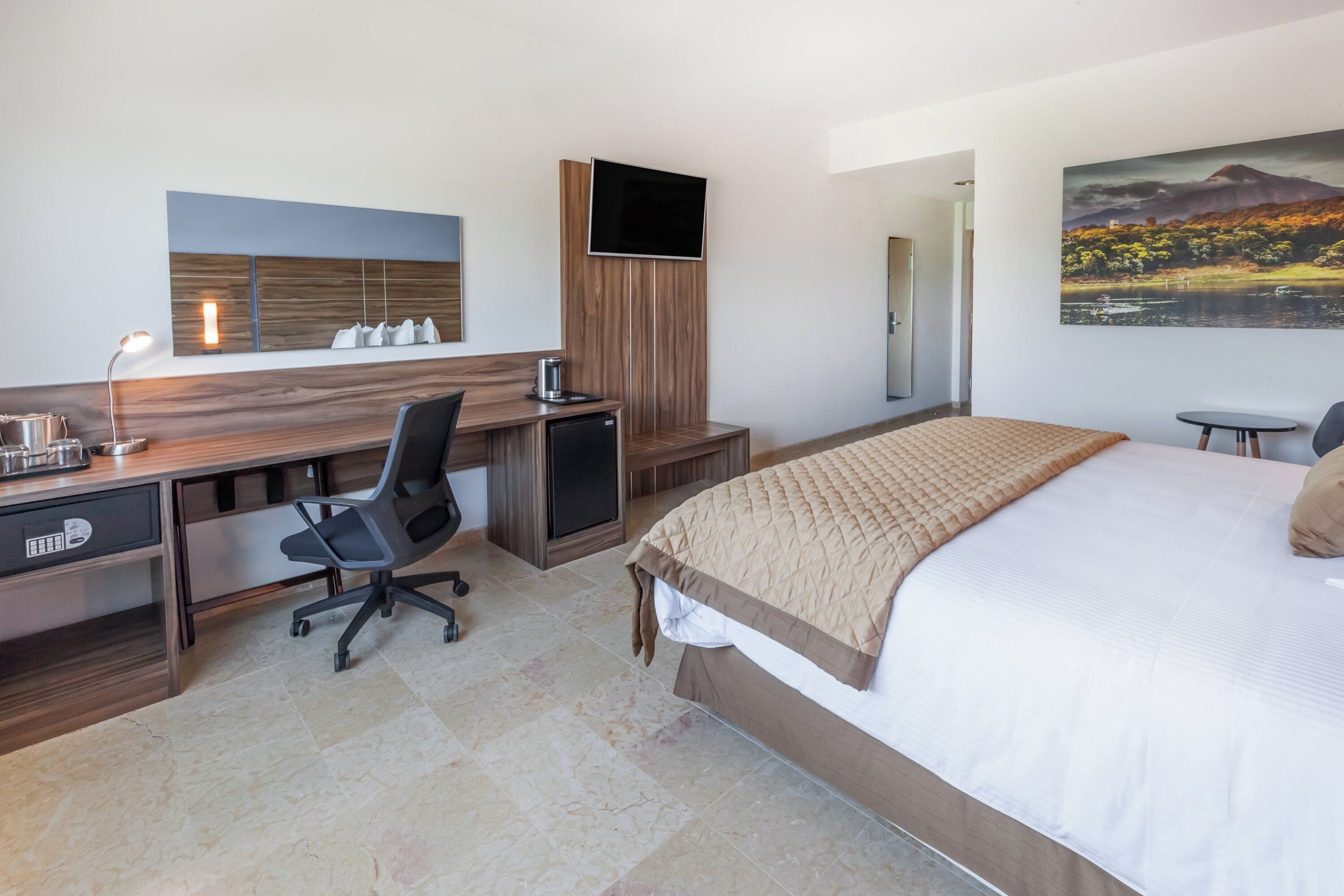 Una habitación de hotel con cama, escritorio, silla, televisor y una obra de arte en la pared.