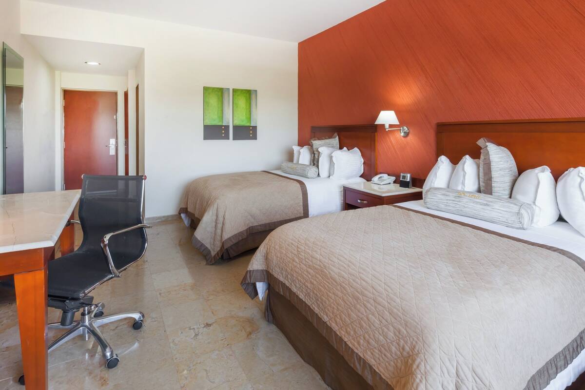 A hotel room with two beds, a desk, a chair, and a nightstand with a lamp.