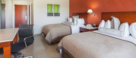 A hotel room with two beds, a desk, a chair, and a nightstand with a lamp.