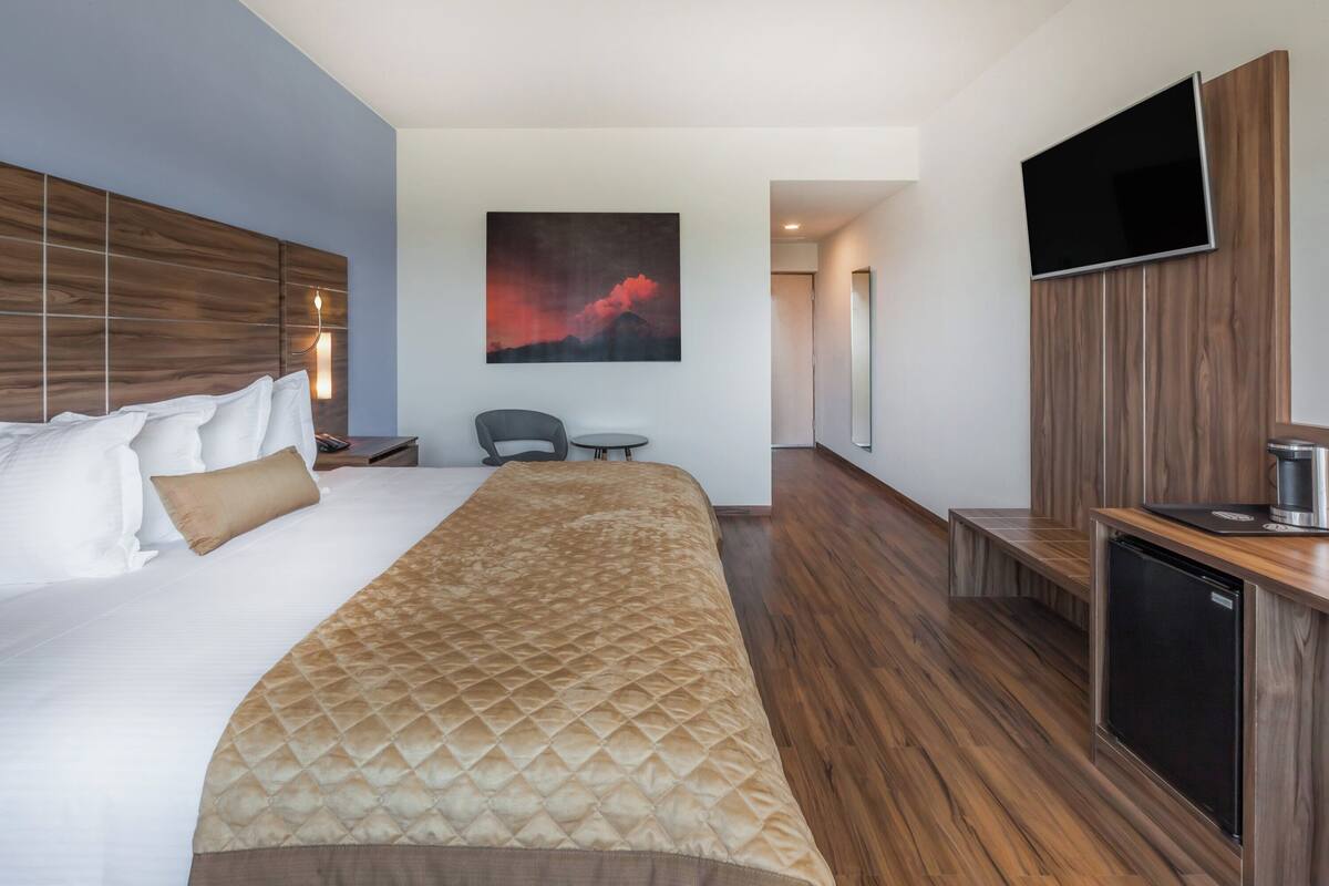 A hotel room with a large bed, a TV mounted on the wall, a small seating area, and a kitchenette.