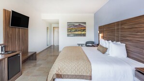 A modern hotel room with a large bed, a flat-screen TV mounted on a wooden panel, a small desk with a chair, and a painting of a landscape on the wall.