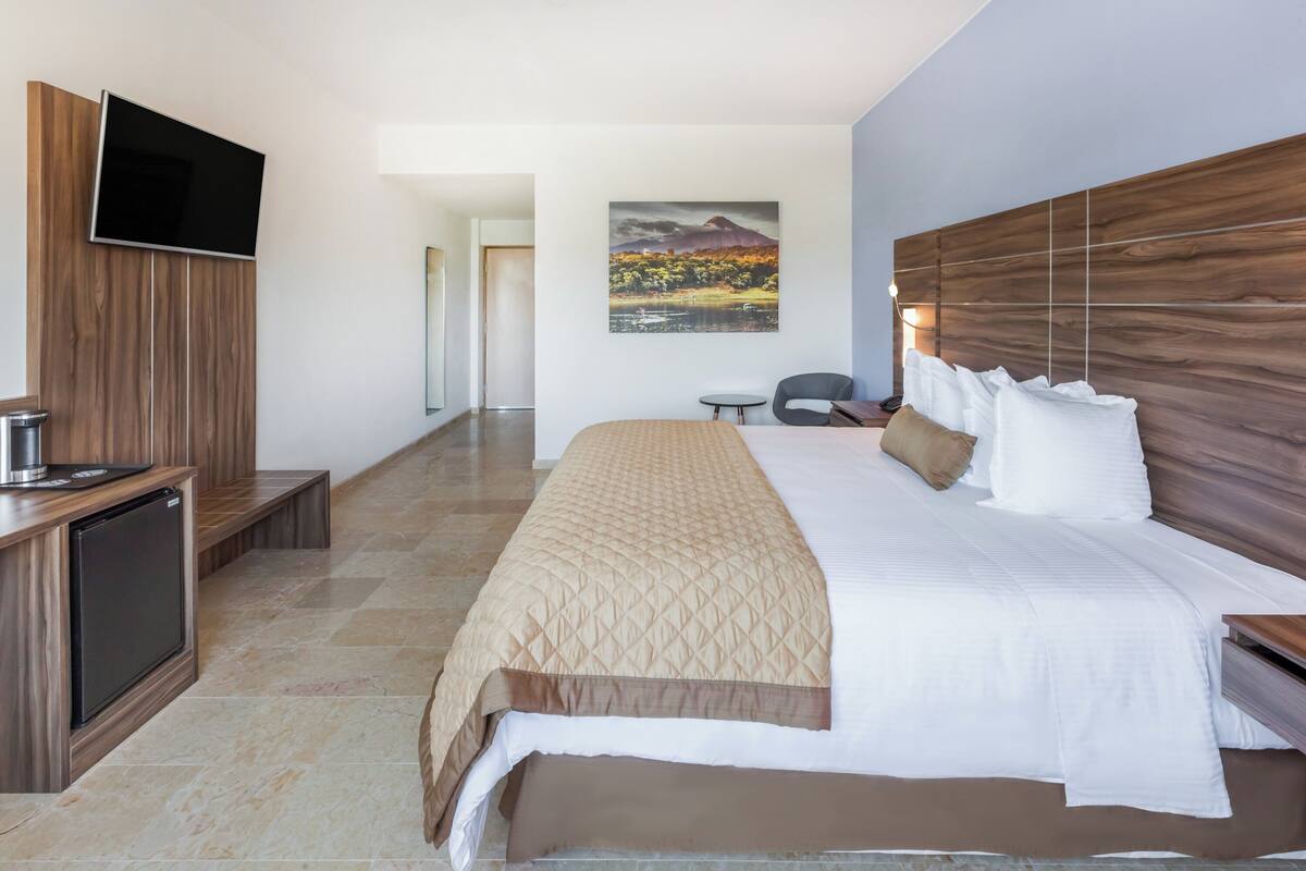 A modern hotel room with a large bed, a flat-screen TV mounted on a wooden panel, a small desk with a chair, and a painting of a landscape on the wall.