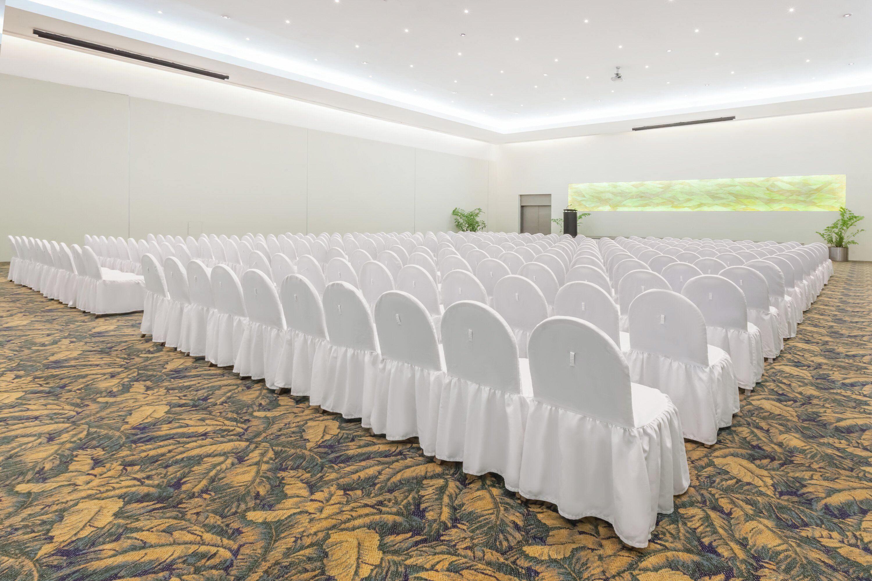 A conference room with rows of white chairs, a carpeted floor, and a large screen on the wall.