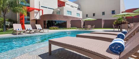Zona de piscina moderna de hotel con sillones, sombrillas y una piscina azul cristalina.
