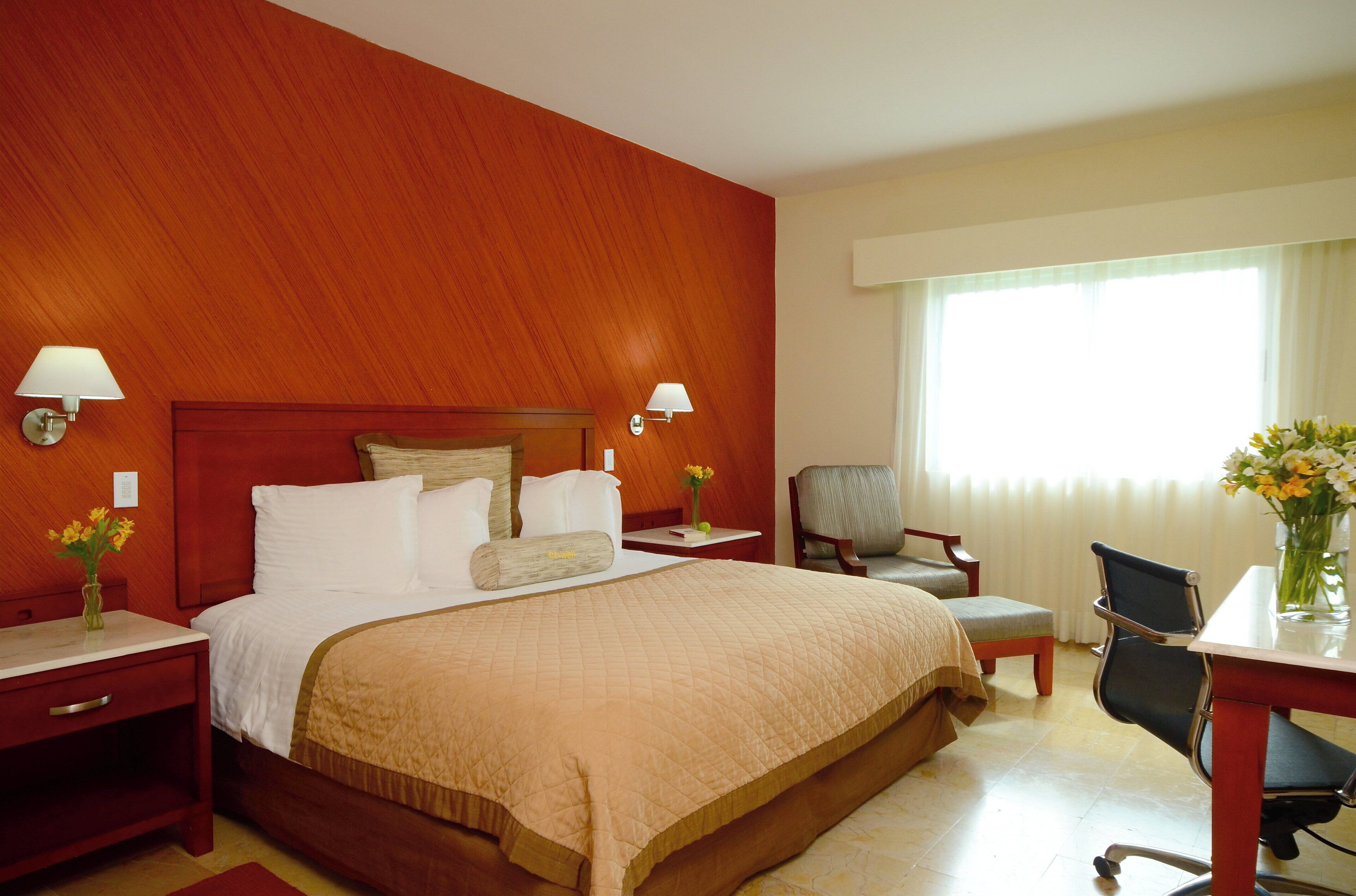 A hotel room with a large bed, two bedside tables with lamps, a desk with a chair, and a window with curtains.