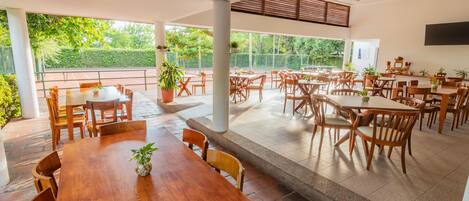 A spacious outdoor dining area with multiple wooden tables and chairs, surrounded by large windows offering views of greenery.
