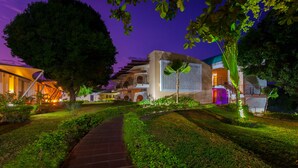 Un sendero bien iluminado que conduce a un edificio moderno, con un gran árbol y abundante vegetación.