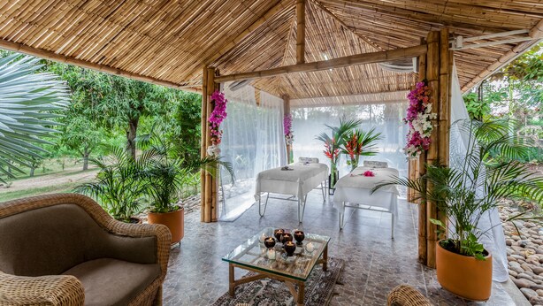 Un área de estar al aire libre techada con muebles de mimbre, una mesa con cubierta de vidrio y velas, y plantas en macetas.