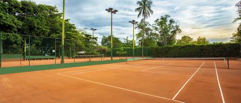 Una cancha de tenis con vallas verdes, luces solares y árboles al fondo.