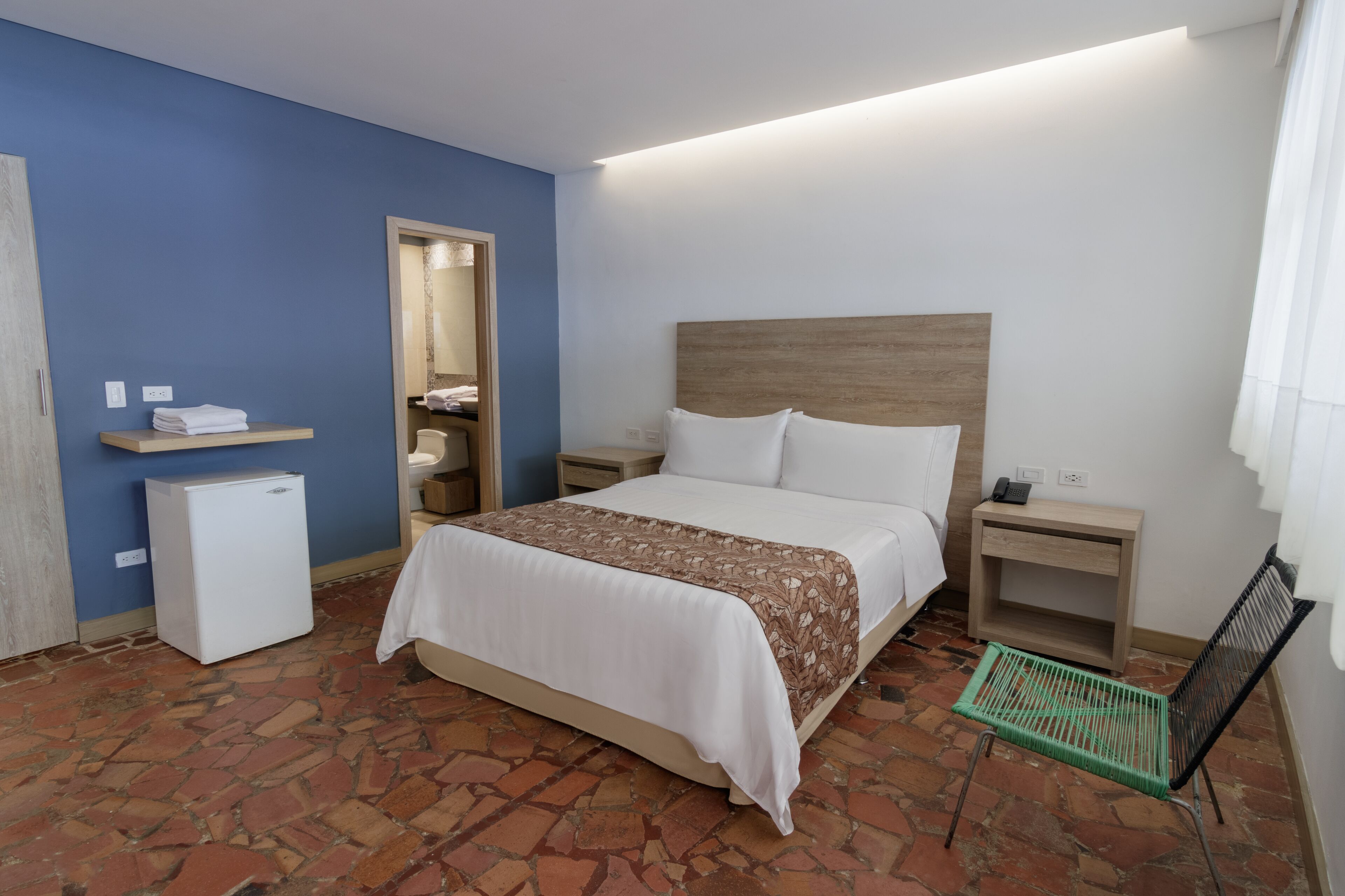 A hotel room with a bed, a small refrigerator, a nightstand, a chair, and a bathroom visible through an open door.