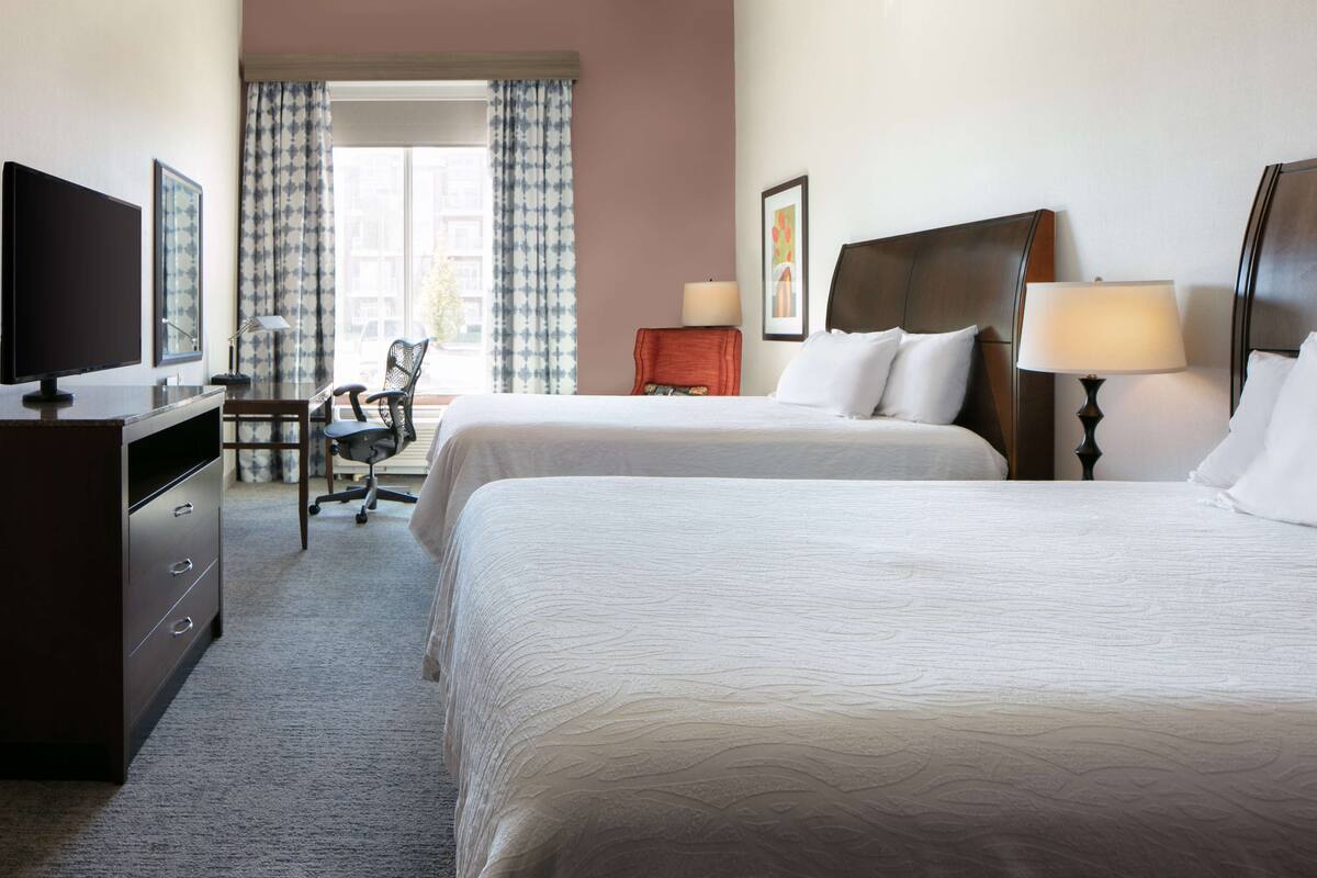 A hotel room with two beds, a desk with a TV, a chair, and a lamp.