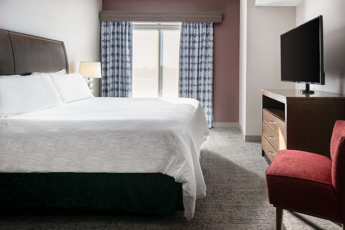 A hotel room with a large bed, a TV, a red chair, and a window with curtains.