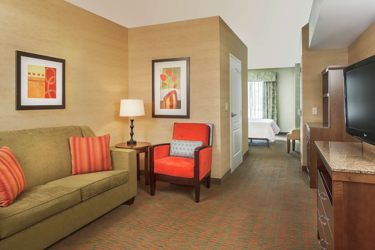 A hotel room with a green sofa, a red armchair, a small table with a lamp, a TV, and artwork on the walls.
