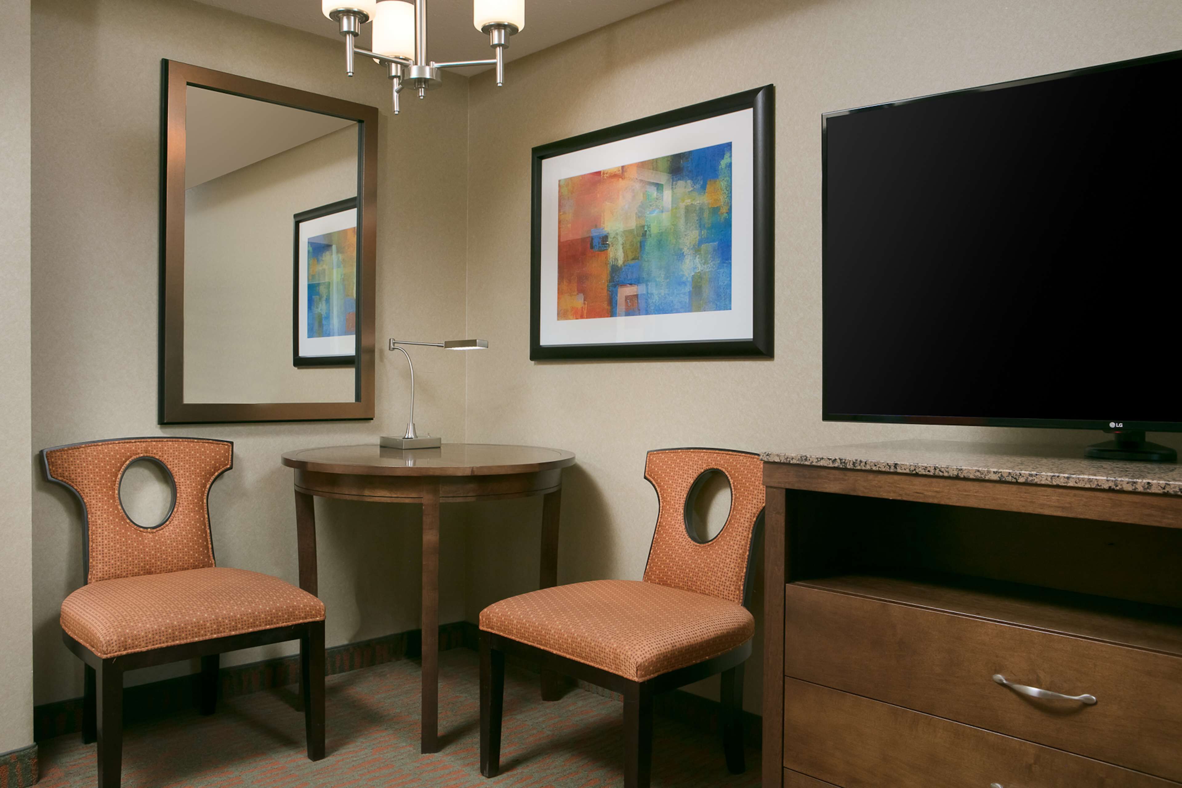 A hotel room with a large mirror, a flat-screen TV, two chairs, a desk, and a framed abstract painting on the wall.