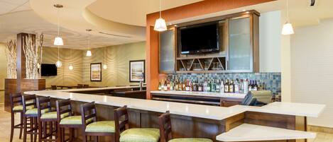 A bar area with a long counter, multiple bar stools, a television mounted above the bar, and a variety of bottles and glasses on the counter.