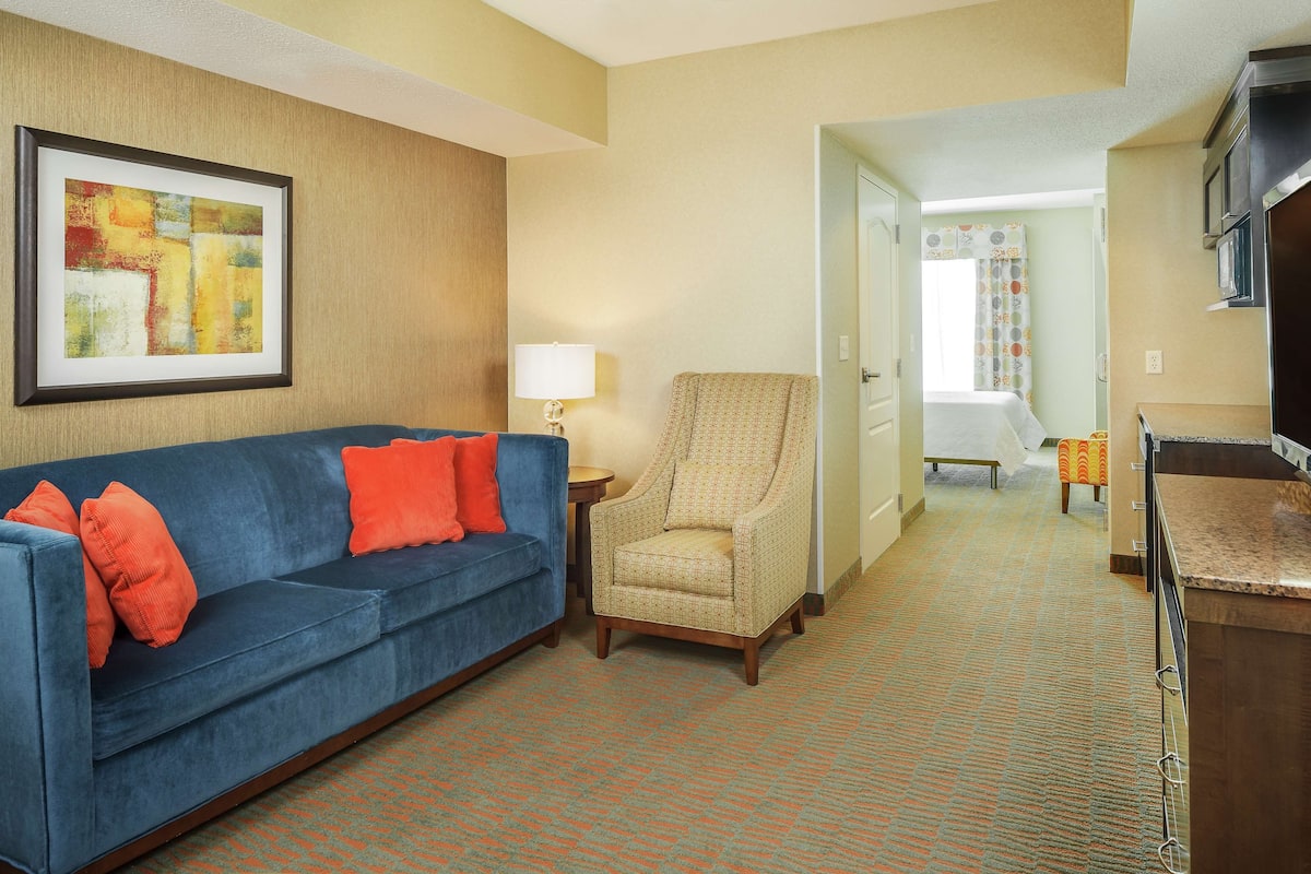 A hotel room with a blue sofa, a patterned armchair, a small table with a lamp, and a kitchen area.