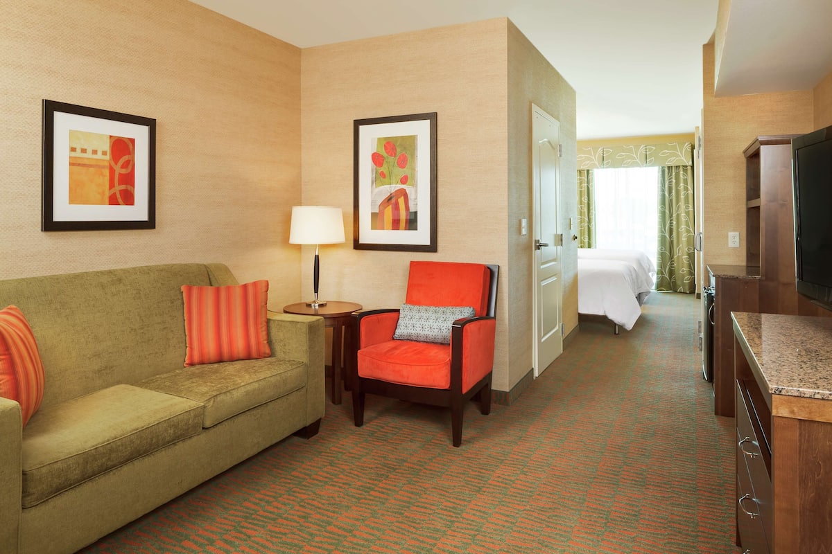 A hotel room with a green sofa, an orange armchair, a small table with a lamp, a bed with a white bedspread, and a television on a wooden stand.