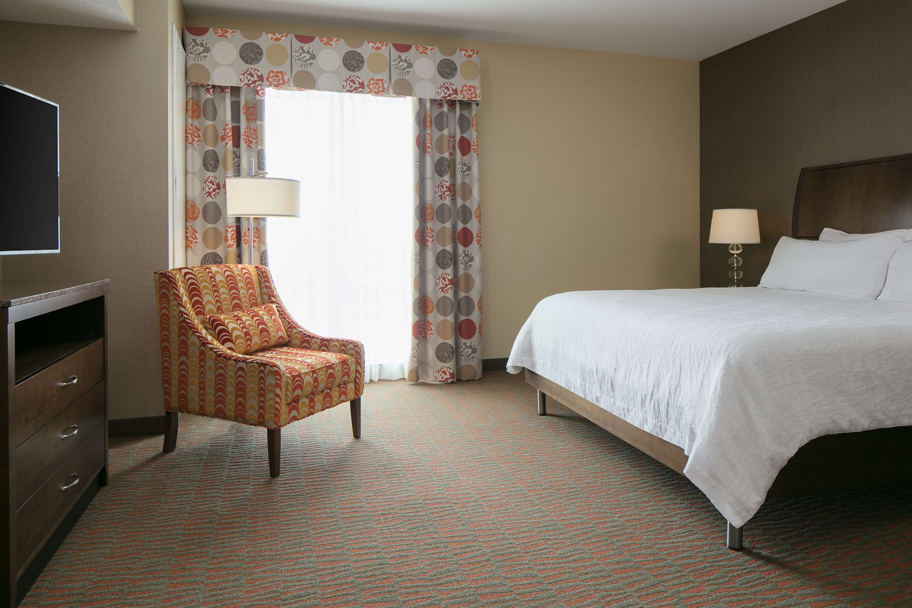 A hotel room with a bed, a chair, a TV, and a lamp.