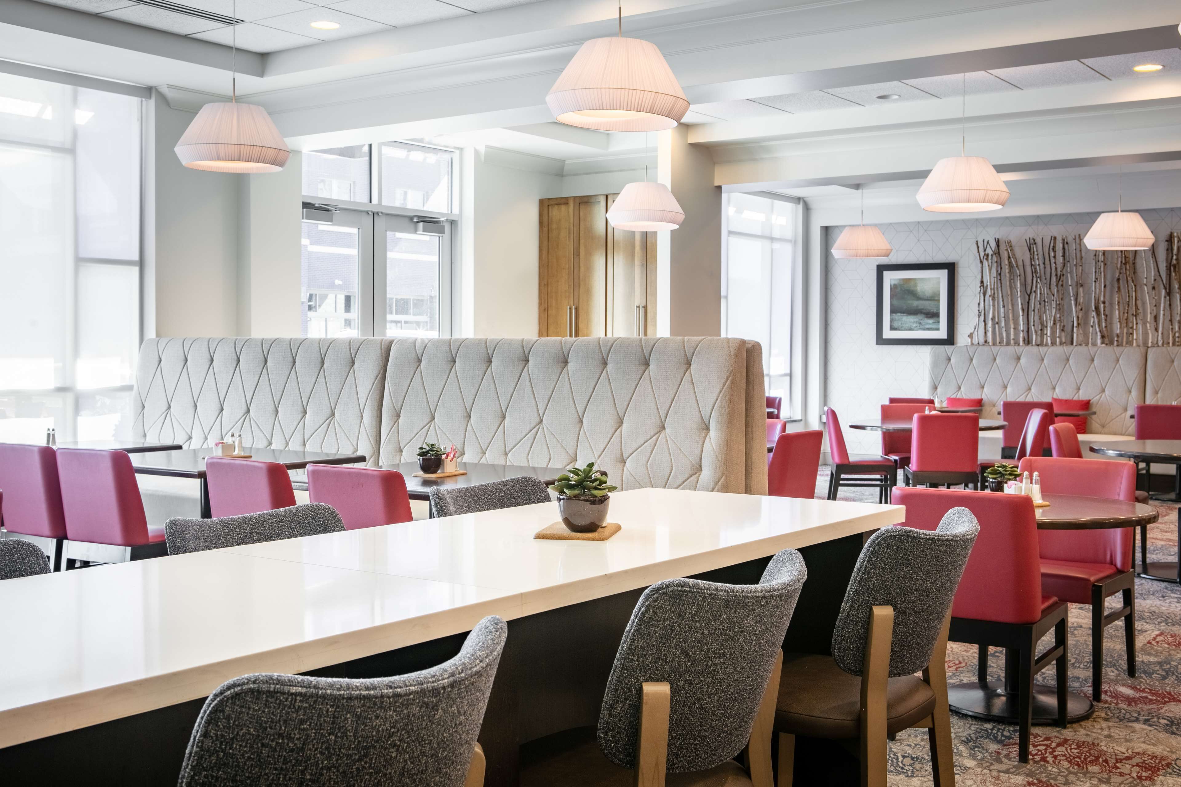Un espace de salle à manger moderne avec une longue table, des chaises à revêtement rouge et gris, et des luminaires suspendus.