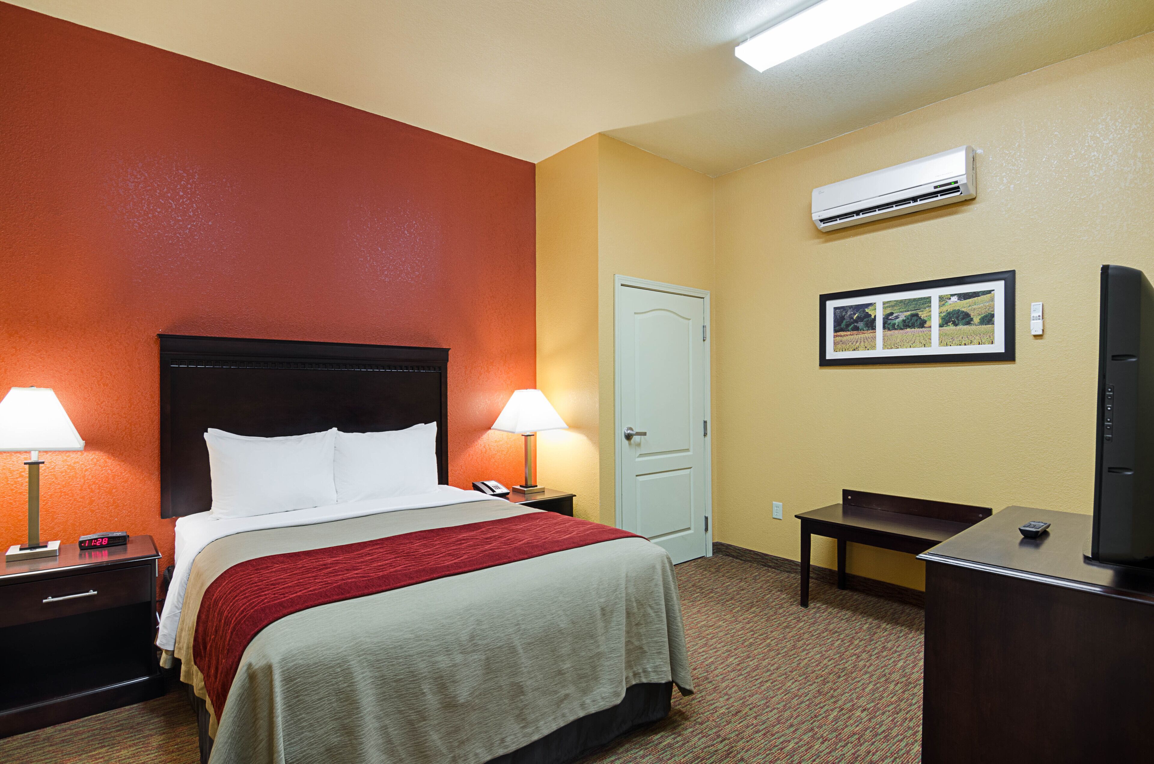 A hotel room with a bed, bedside lamps, a desk, a television, and an air conditioning unit.