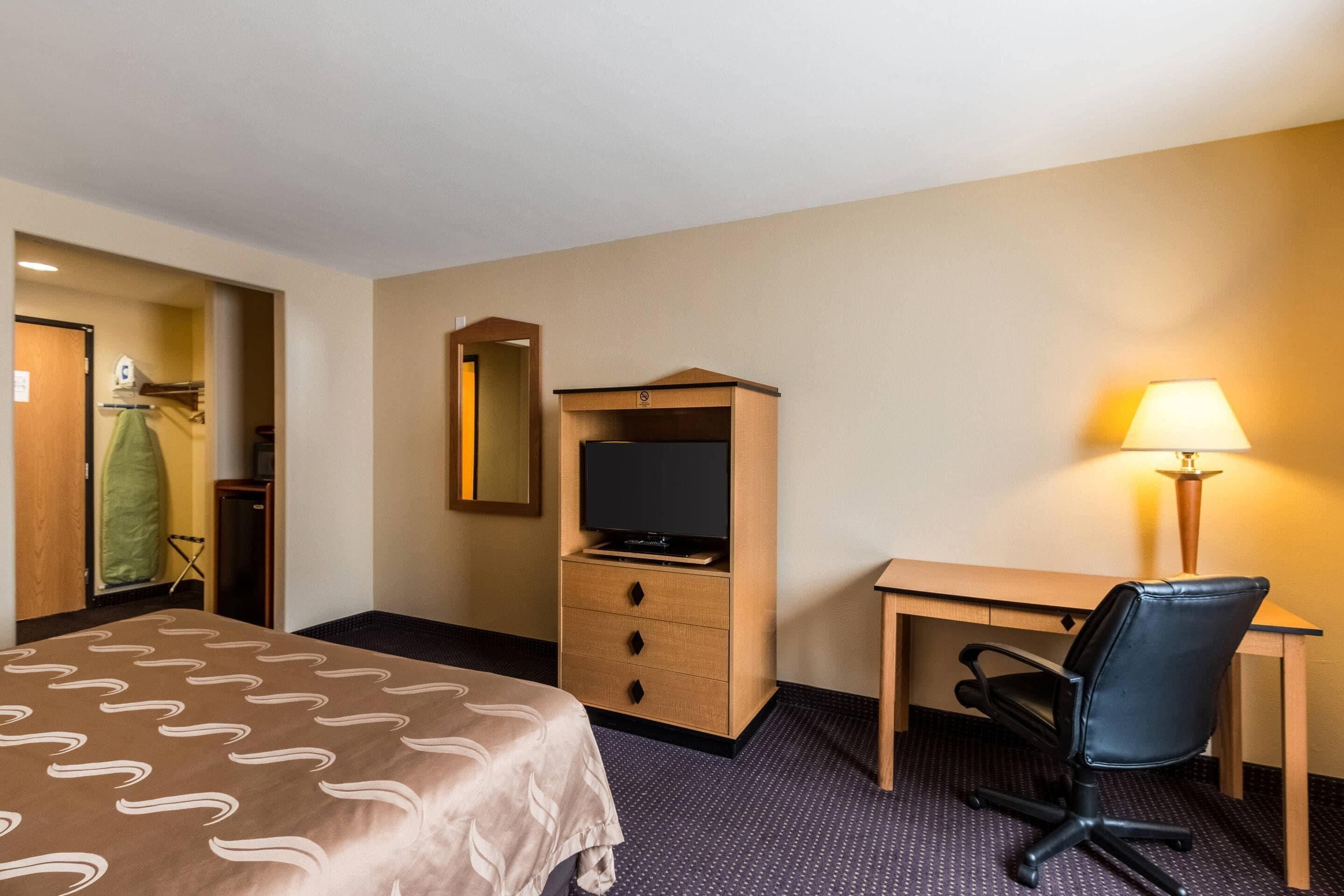 A hotel room with a bed, a desk with a chair, a television, a lamp, and a wardrobe.