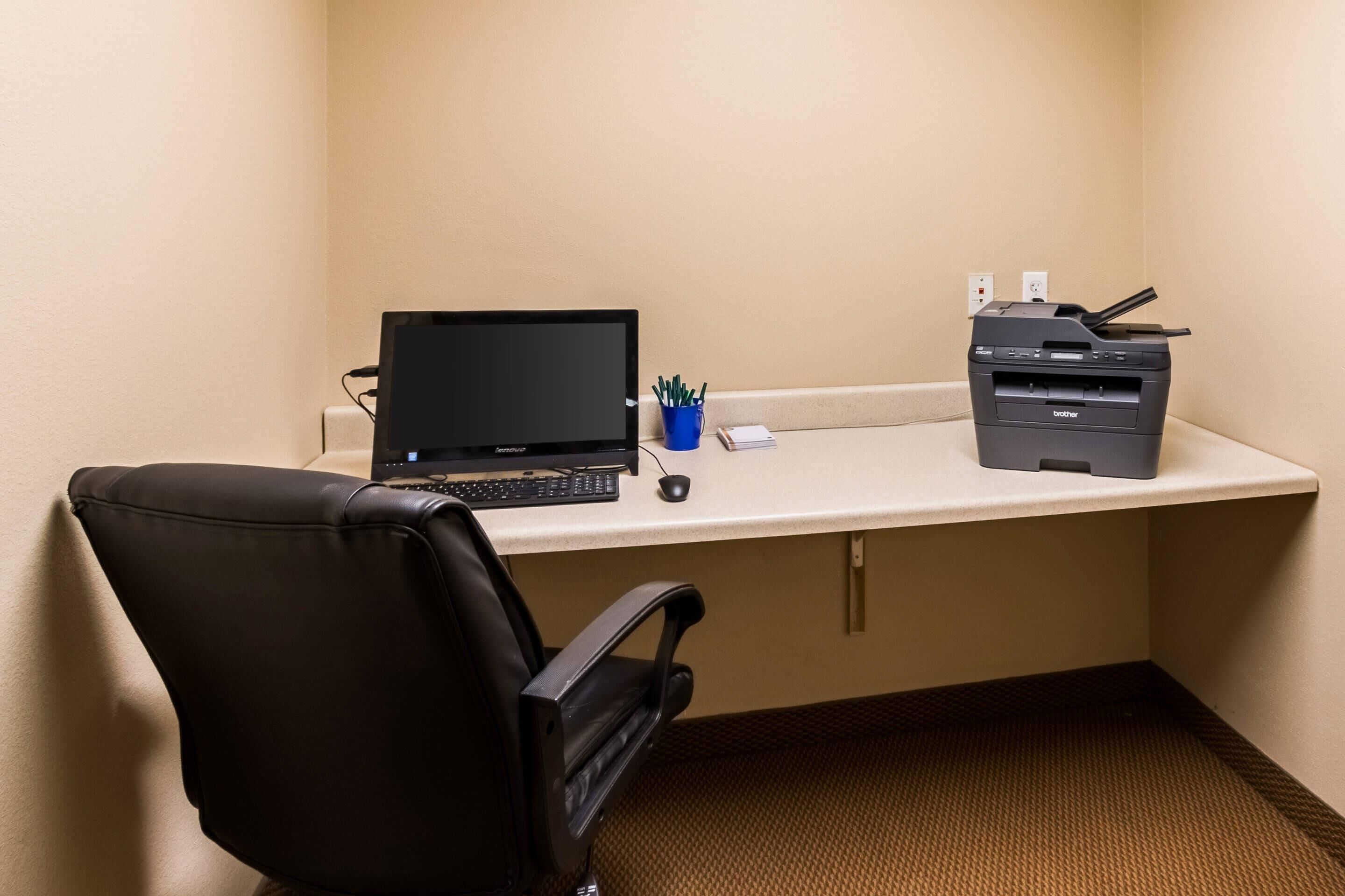 A hotel office with a desk, laptop, printer, chair, and a small blue container with pens.
