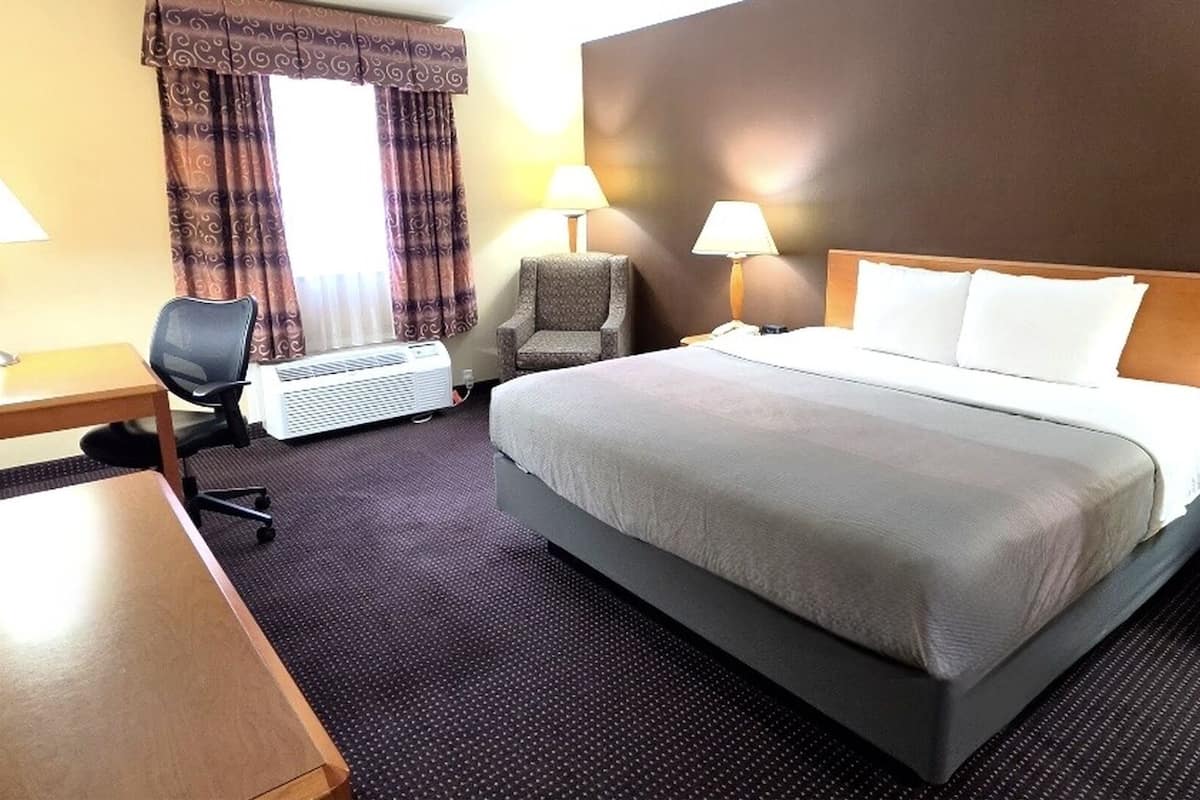 A hotel room with a bed, two bedside lamps, a desk, a chair, and an air conditioning unit.