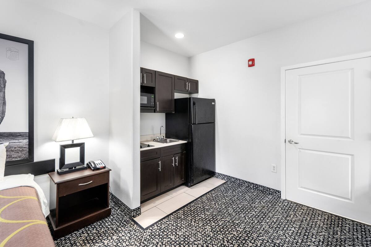 A compact hotel room with a kitchenette, a bed, a bedside table with a lamp, and a framed picture on the wall.