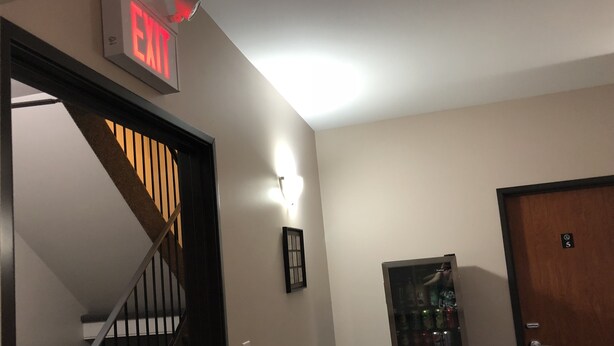 A hallway with a staircase, an exit sign, a refrigerator, and a door. - Budget Lodge (Saskatoon)