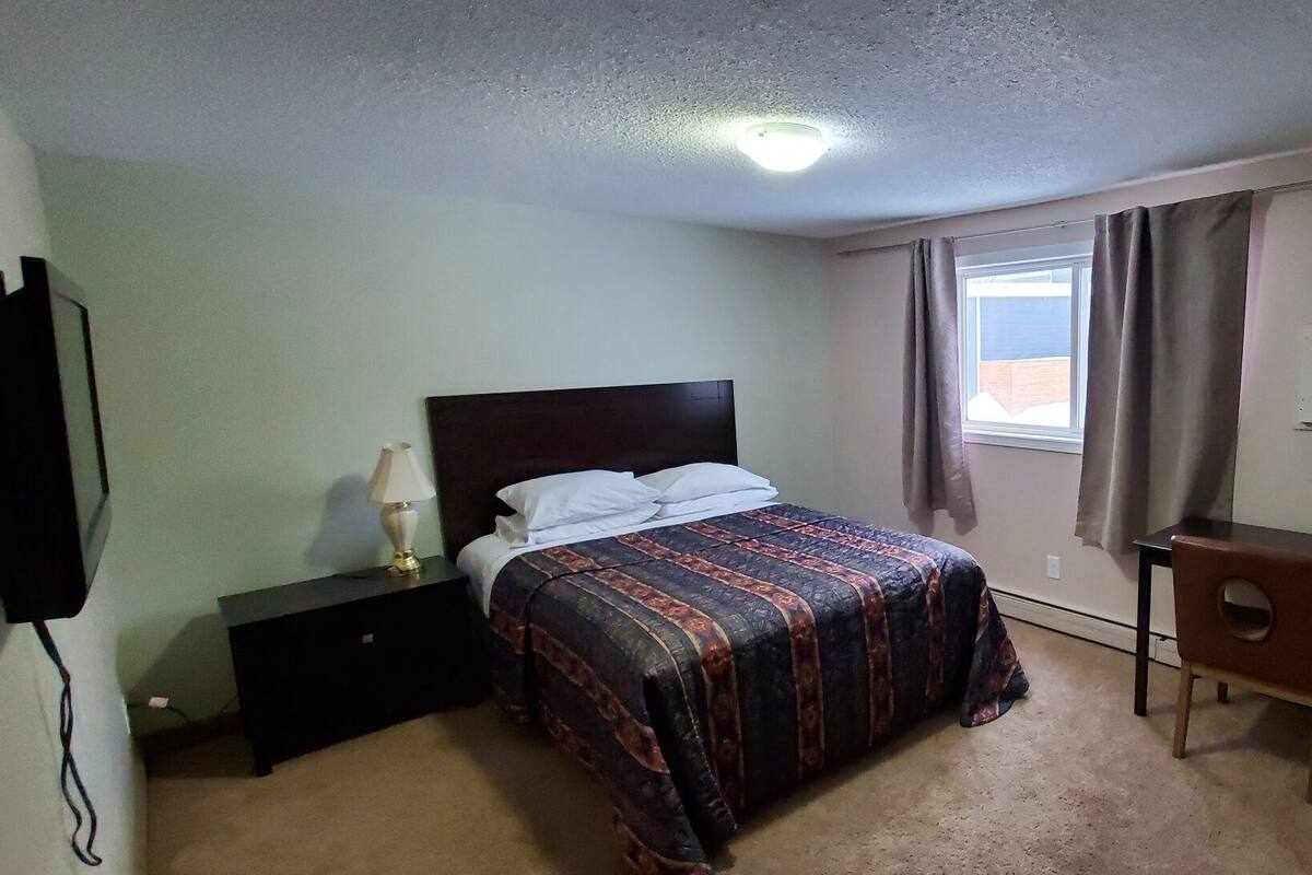 A bedroom with a bed, a desk, a chair, a TV, and a window with curtains.