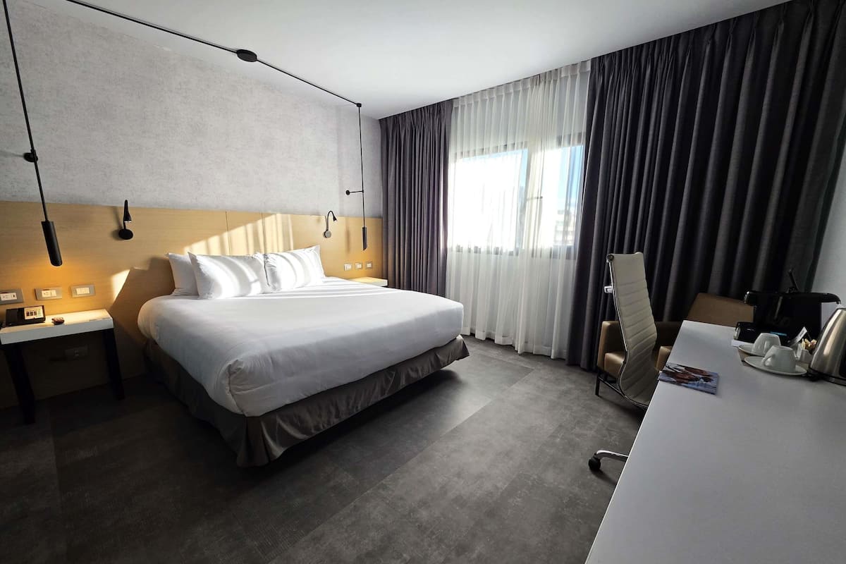 A hotel room with a bed, bedside tables, a desk with a chair, a coffee maker, and a window with curtains.