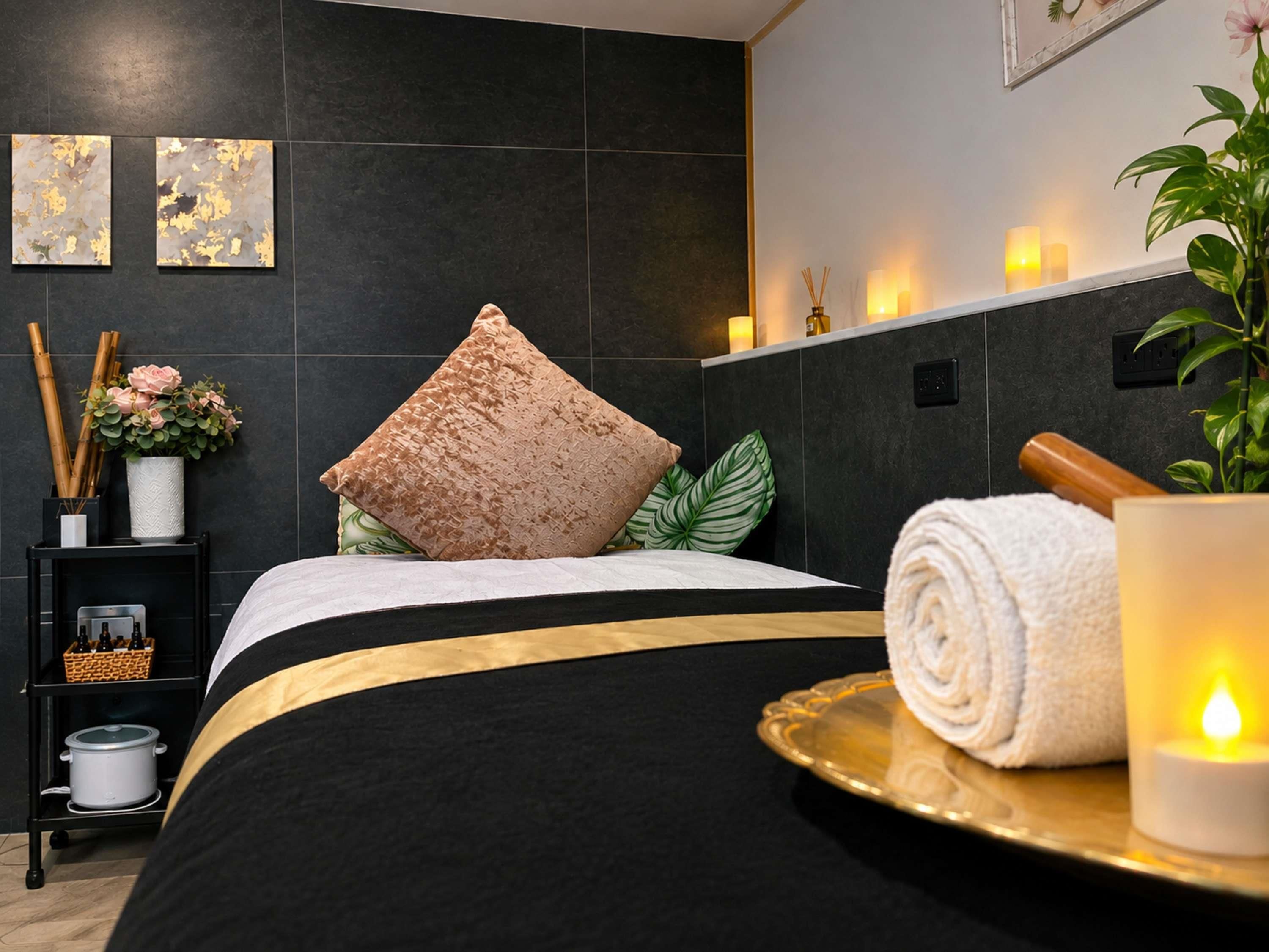 Un dormitorio moderno con colcha negra y dorada, una almohada decorativa, una mesita de noche con una planta pequeña y velas, y una bandeja con una toalla enrollada y una vela encendida.