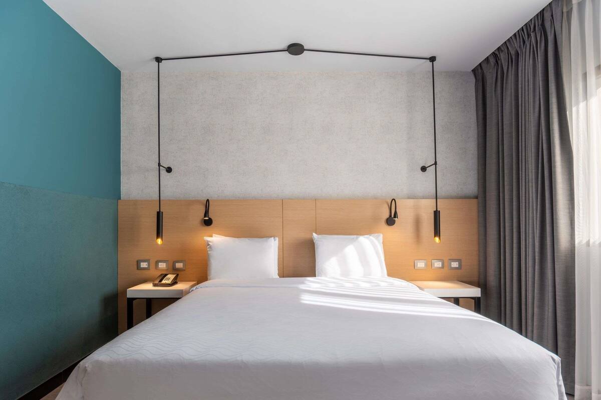 A modern hotel room with a large bed, two bedside tables, a wooden headboard, and pendant lights.