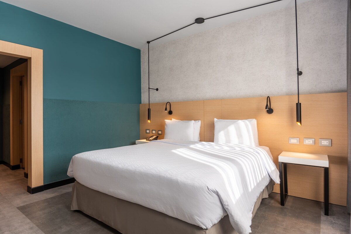 A modern hotel room with a large bed, bedside tables, and a built-in desk.