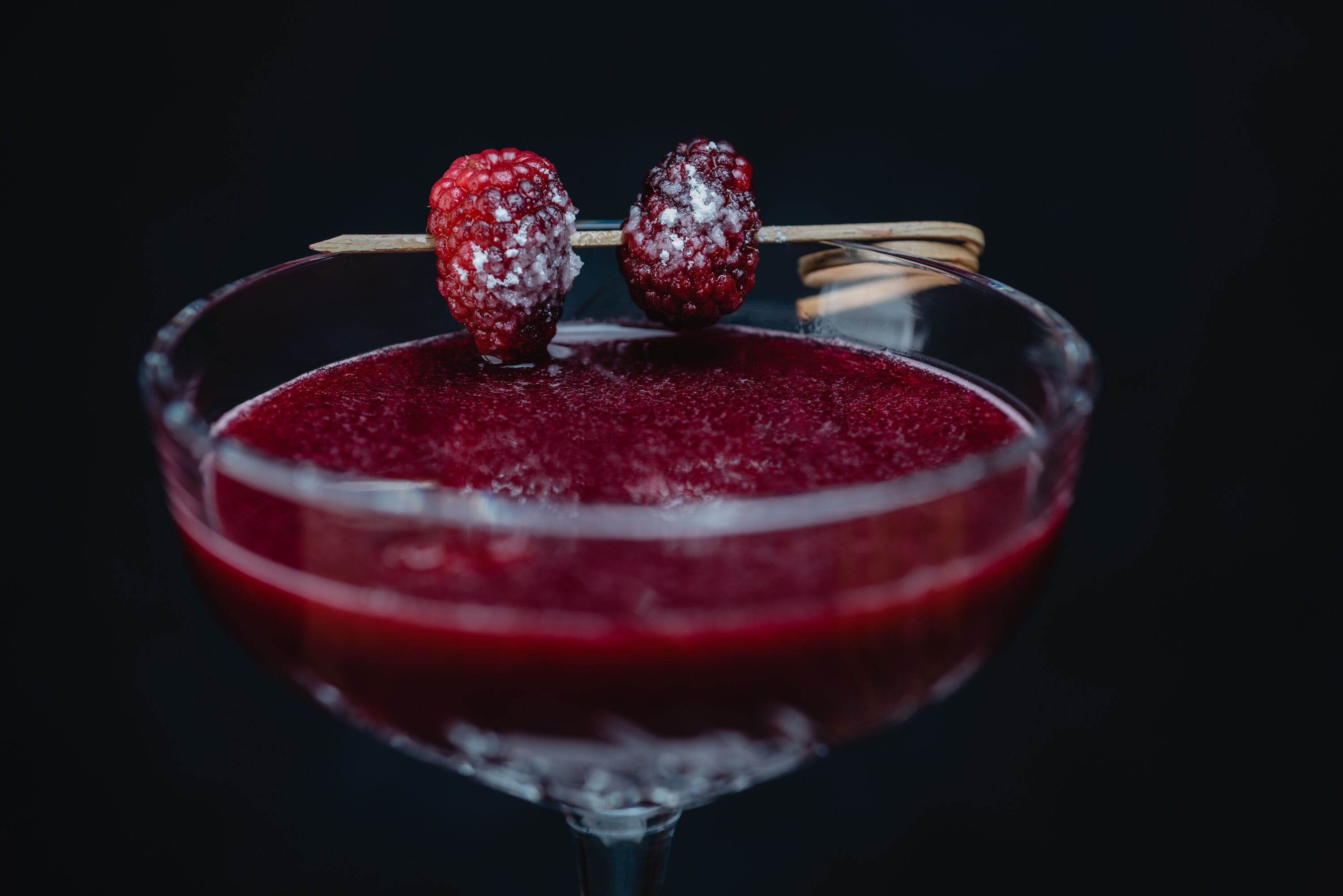 Una copa de martini con una bebida rojo oscuro y dos frambuesas ensartadas en un palillo.