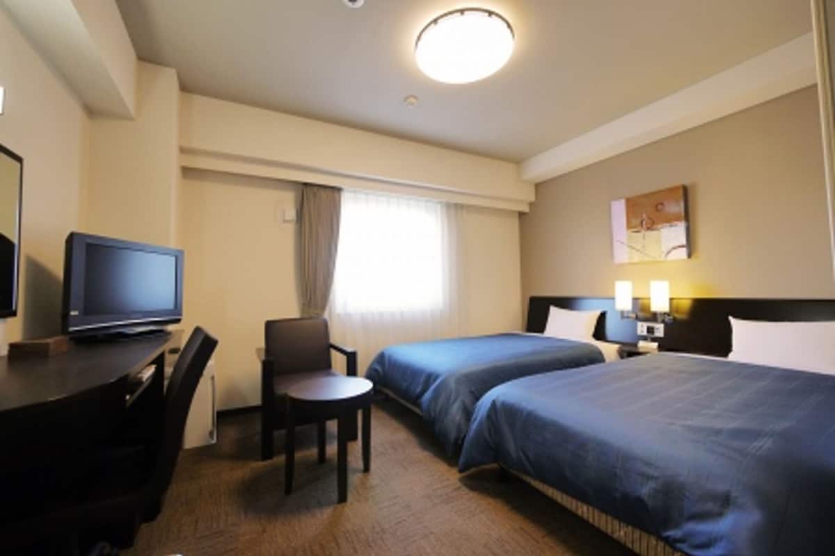 A hotel room with two beds, a desk with a television, a chair, and a nightstand with lamps.