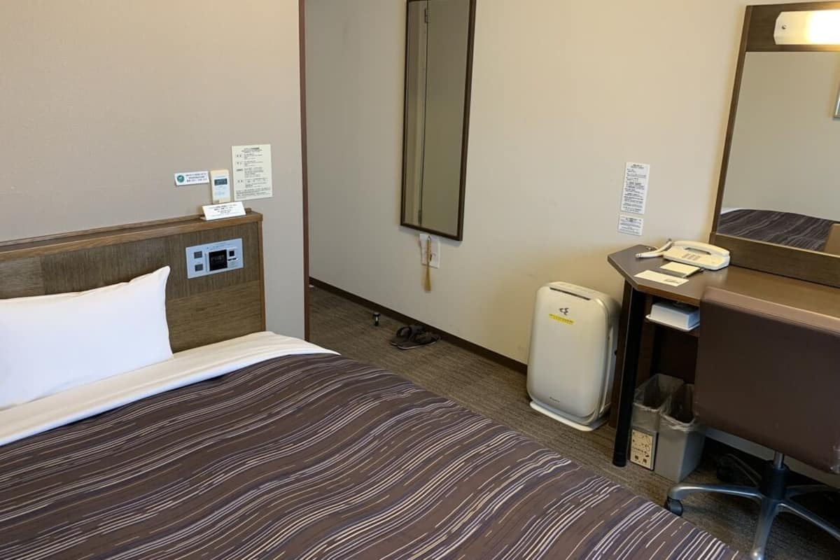 A hotel room with a bed, a desk, a chair, a mirror, a phone, and a trash bin.